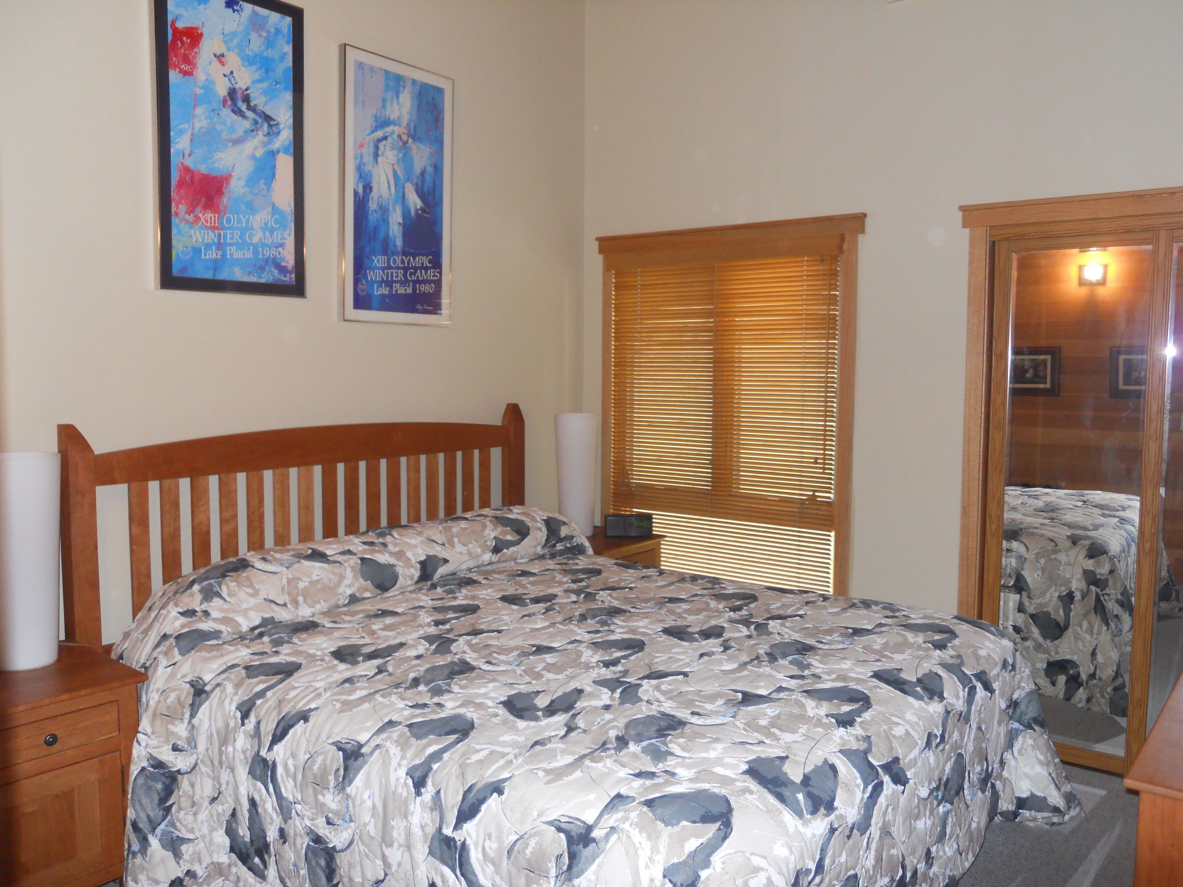 A bedroom with a wooden bed frame, a large bedspread, a window with blinds, and two posters on the wall.