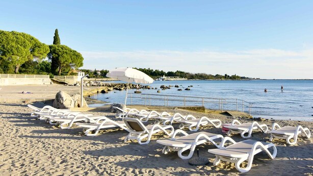 Ein Strand mit weißen Liegestühlen und Sonnenschirmen, einer felsigen Küstenlinie und einem klaren Himmel.