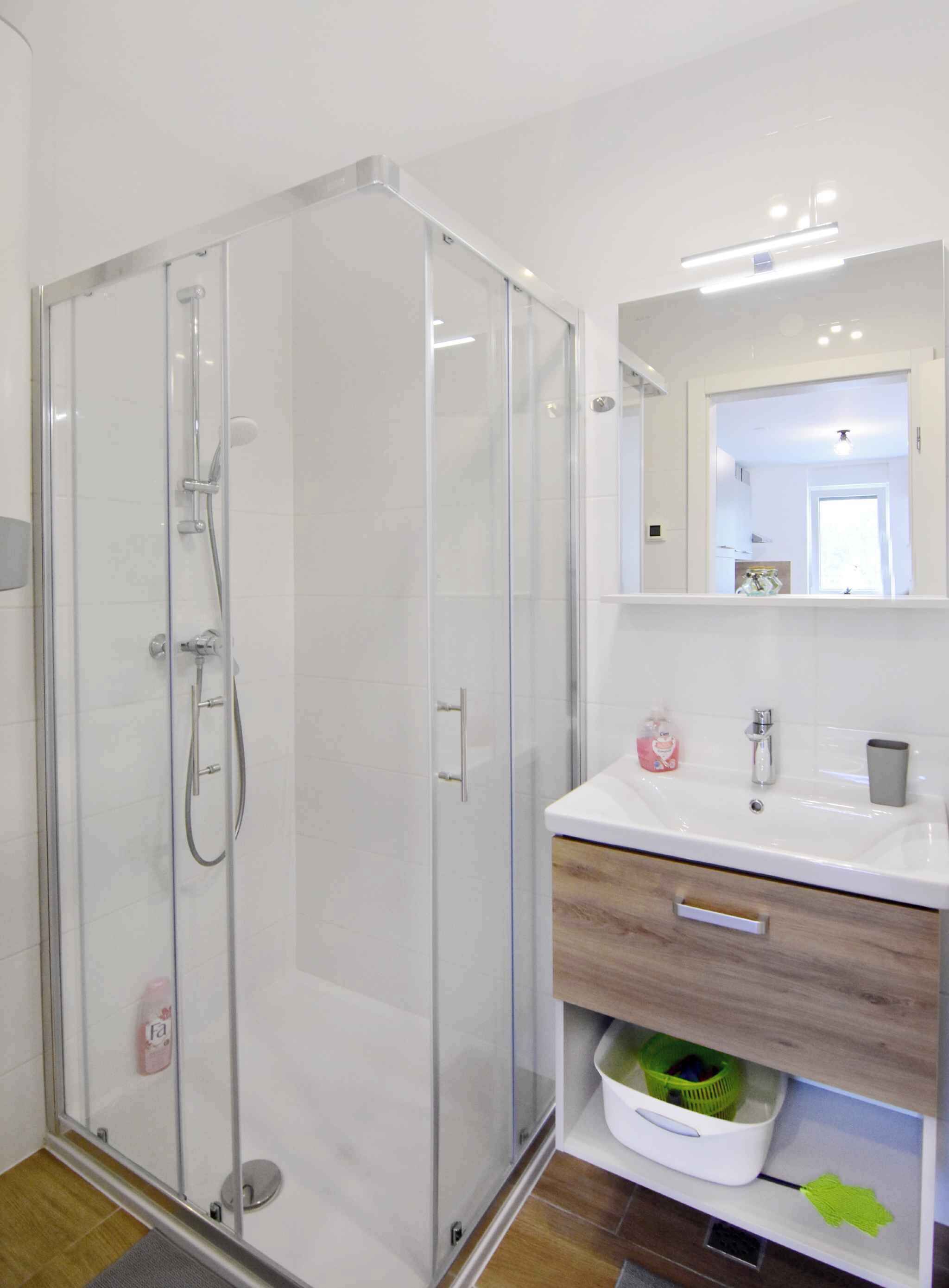 Un baño moderno con ducha envuelta en vidrio, un lavabo blanco con espejo y un mueble de tocador de madera con cajones.