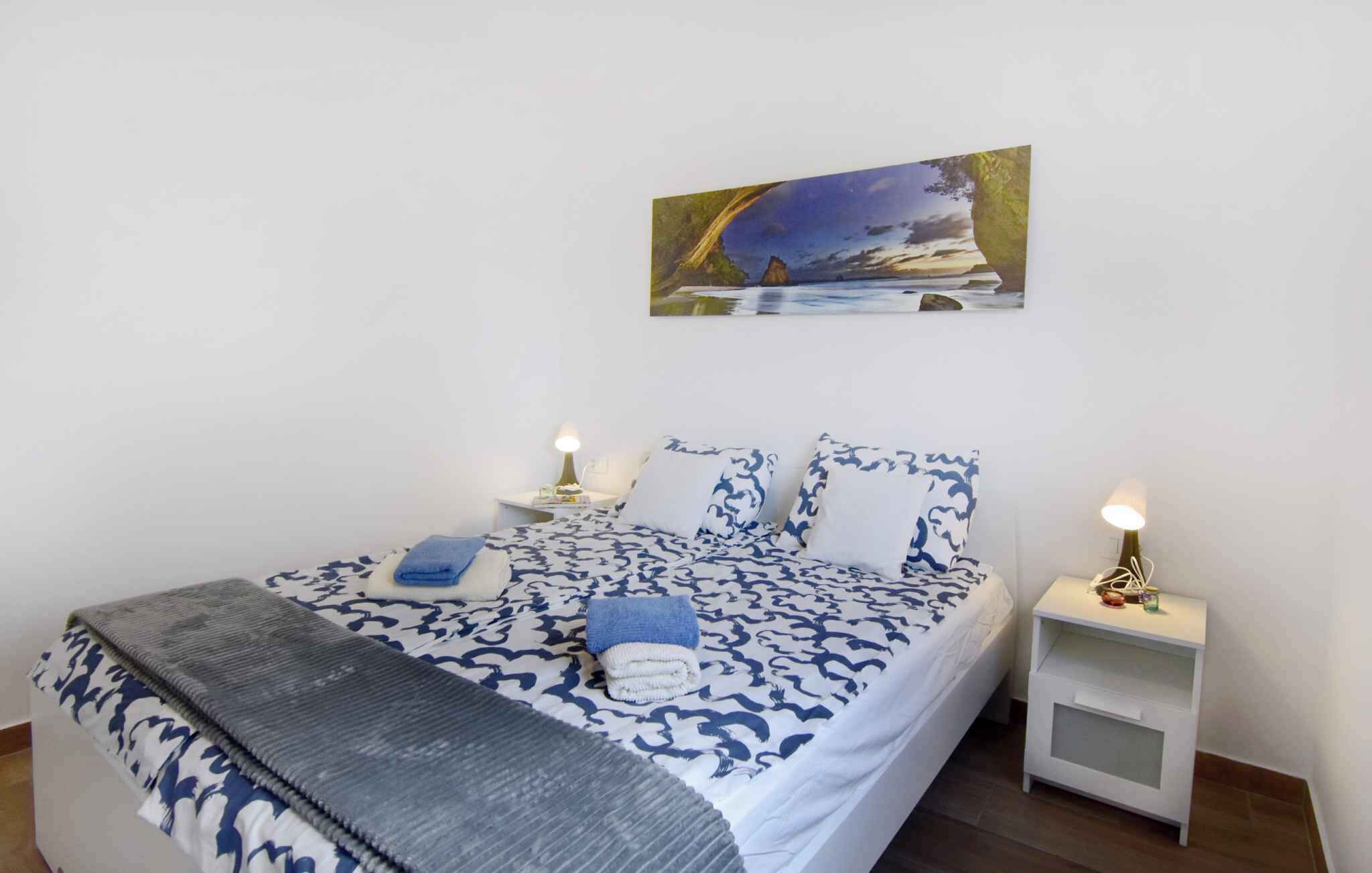 A neatly made bed with a patterned bedspread, two bedside tables with lamps, and a painting of a landscape on the wall.