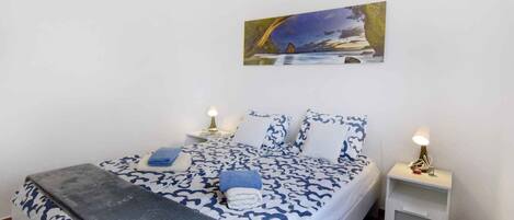 A neatly made bed with a patterned bedspread, two bedside tables with lamps, and a painting of a landscape on the wall.