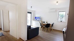 Ein modernes Wohnzimmer mit einer Couch, einem Esstisch und Stühlen. Ein Fernseher steht auf einer Kommode, und ein Fenster bietet Blick auf Grünfläche.