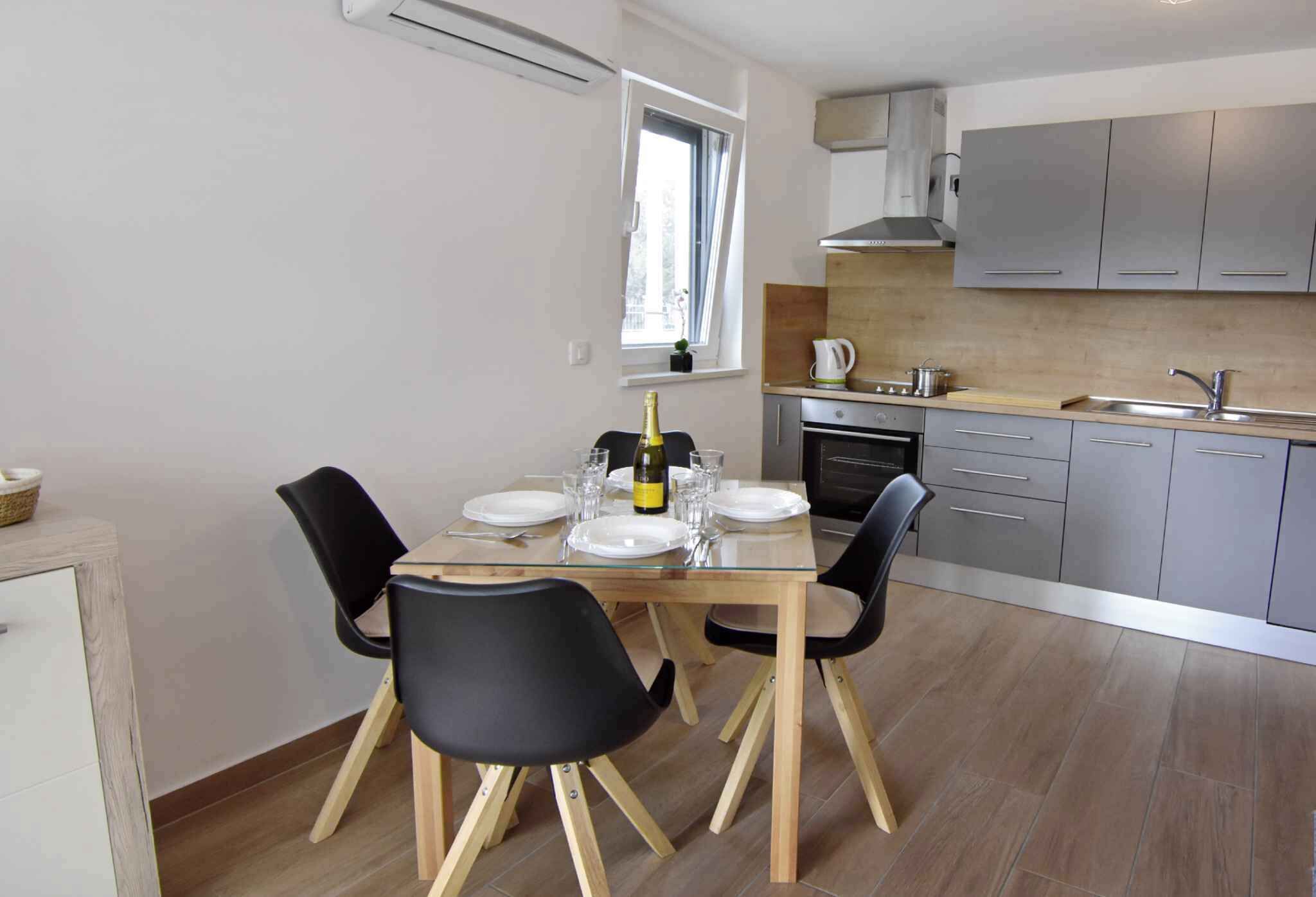 Une cuisine moderne avec des meubles gris, un évier en acier inoxydable et un coin repas comprenant une table en verre et des chaises noires.