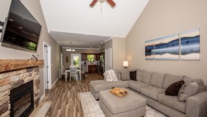A modern living room with a stone fireplace, a grey sectional sofa, a flat-screen TV mounted on the wall, and a ceiling fan. - It is Springtime in TN- comfortable, clean, in a gated community & near food! (Townsend)