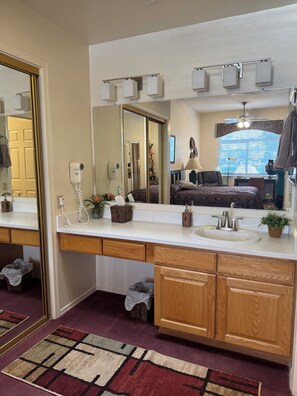 A modern bathroom with a large mirror, a white sink, wooden cabinets, and a patterned rug. - Comfy Studio in SW resort near golf courses and mountain/desert hiking (Green Valley)