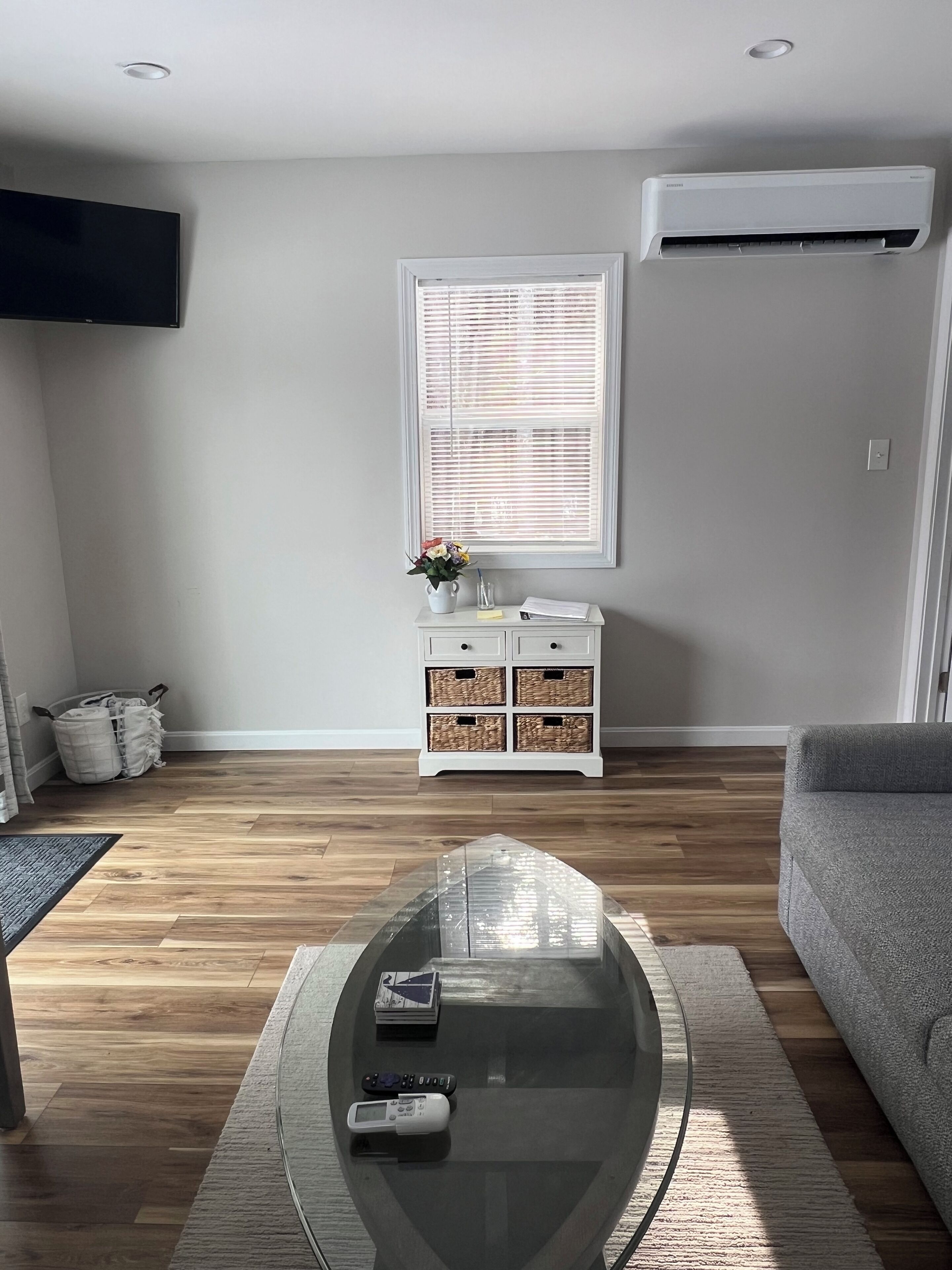 A living room with a glass coffee table, a grey sofa, a white side table with baskets, a window with blinds, and an air conditioning unit.