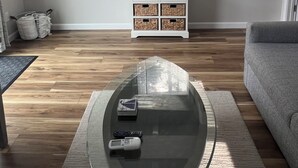 A living room with a glass coffee table, a grey sofa, a white side table with baskets, a window with blinds, and an air conditioning unit. - Cozy quiet Cottage (Deer Isle)
