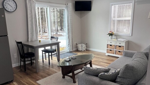 A modern living room with a grey sofa, a dining table with chairs, a glass-top coffee table, a flat-screen TV mounted on the wall, and a large window with blinds. - Cozy quiet Cottage (Deer Isle)