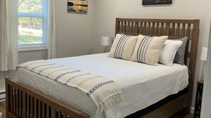 A neatly arranged bedroom with a wooden bed frame, a large window with curtains, and two wall-mounted paintings. - Cozy quiet Cottage (Deer Isle)