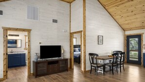 A spacious living area with a wooden ceiling, a dining table with chairs, a TV, and a kitchen area with cabinets and a sink. - The Armadillo Cabin | Great Back Porch (Fredericksburg)