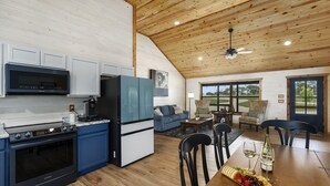 A modern kitchen with a built-in microwave, stove, and refrigerator. There is a dining area with a wooden table, chairs, and a fruit platter. - The Armadillo Cabin | Great Back Porch (Fredericksburg)