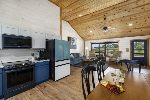 A modern kitchen with a built-in microwave, stove, and refrigerator. There is a dining area with a wooden table, chairs, and a fruit platter. - The Armadillo Cabin | Great Back Porch (Fredericksburg)