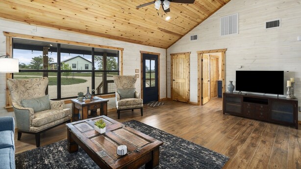 A spacious living room with a high ceiling, wooden walls, and large windows. There is a television, a coffee table, and two sofas. - The Armadillo Cabin | Great Back Porch (Fredericksburg)