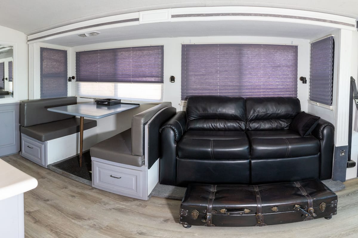 A compact RV interior with a kitchenette, a dining area, a seating area with a black leather sofa, and a small table.