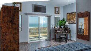 A bedroom with a large bed, a desk with a lamp, a TV mounted on the wall, and a sliding glass door leading to a balcony with a view of the beach. - El Capitan Resort (Playas de Rosarito)