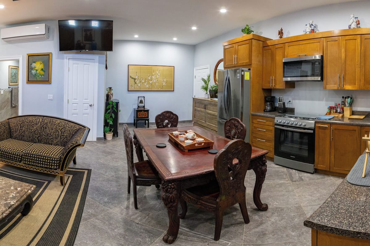 A spacious living room with a sofa, coffee table, and a dining area with a wooden table and chairs. A kitchen with wooden cabinets and modern appliances is visible.