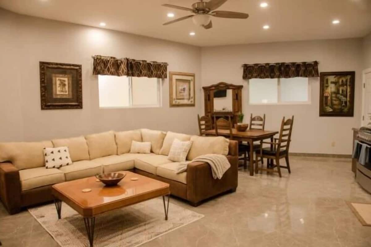 A living room with a sectional sofa, a coffee table, a dining table with chairs, a ceiling fan, and a kitchen area with a stove.