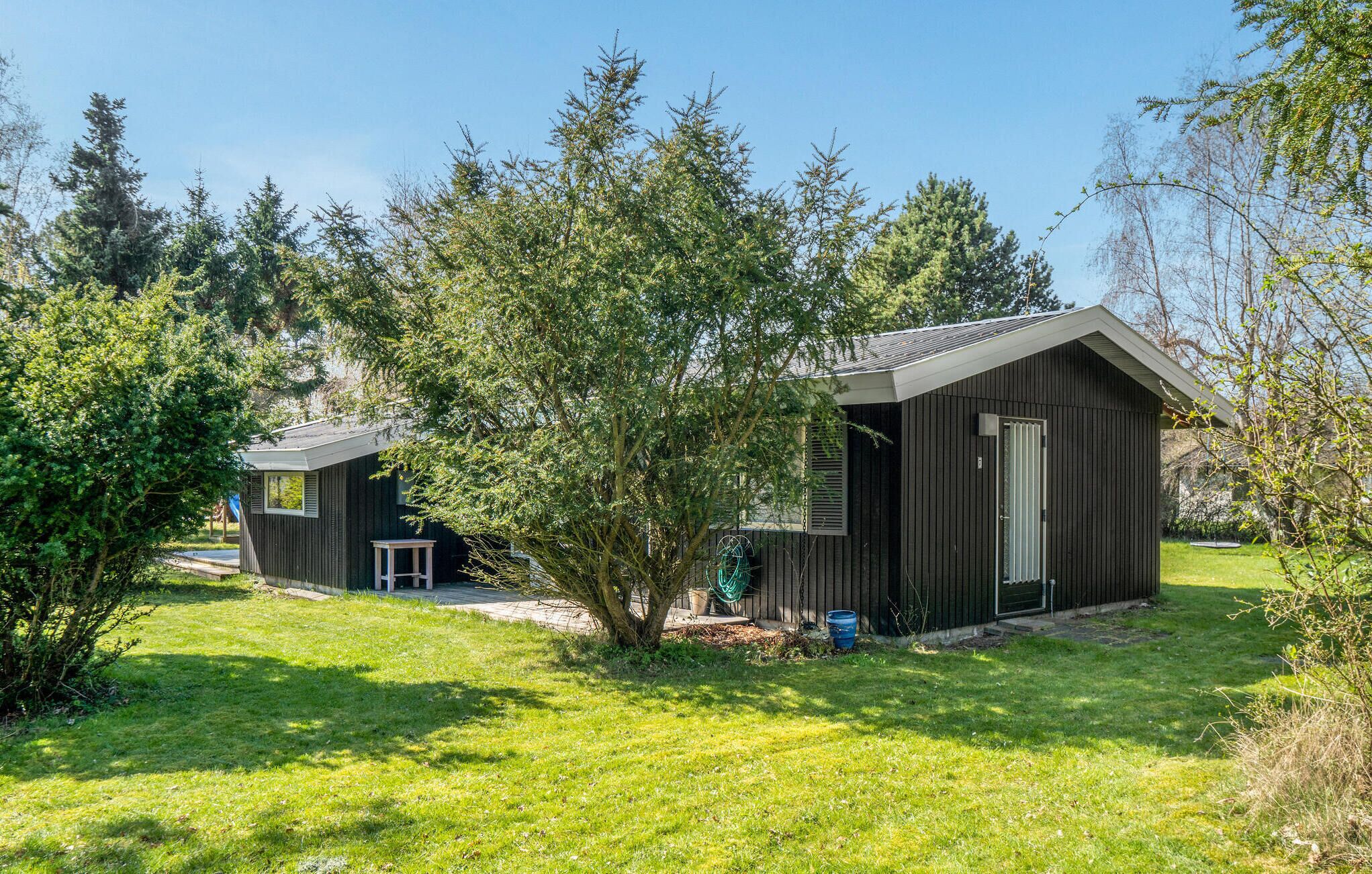 A small, dark wooden cabin with a sloped roof, surrounded by lush green grass and trees. - 3 bedroom lovely home in Sjællands Odde (Sjællands Odde)