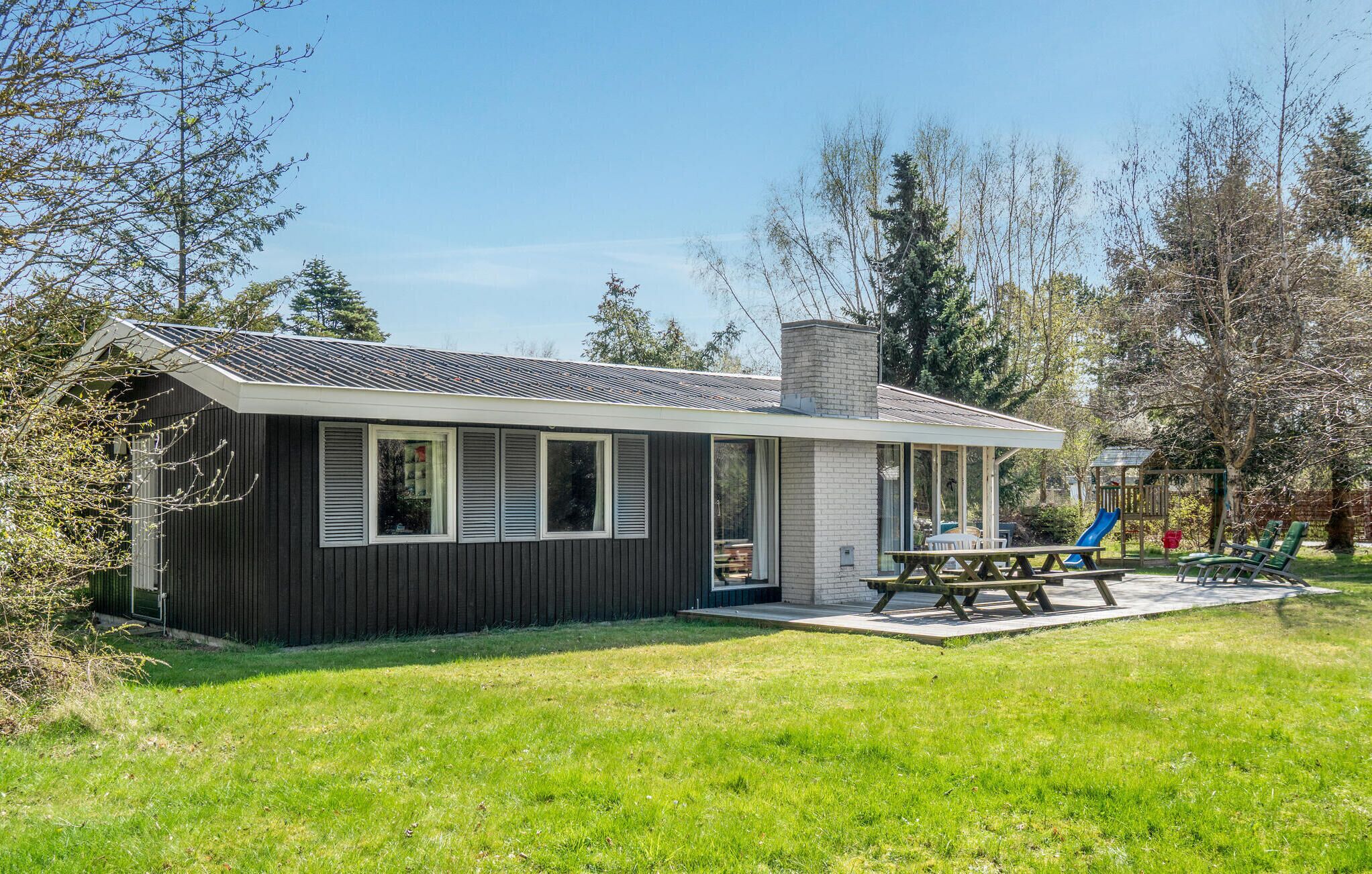 Et etages hus med mørk facade, skorsten og overdækket terrasse med et bord og stole.