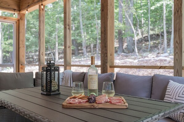 Una mesa de madera con una botella de vino, copas y una tabla de embutidos y queso, ubicada en una terraza de madera con vista a árboles.