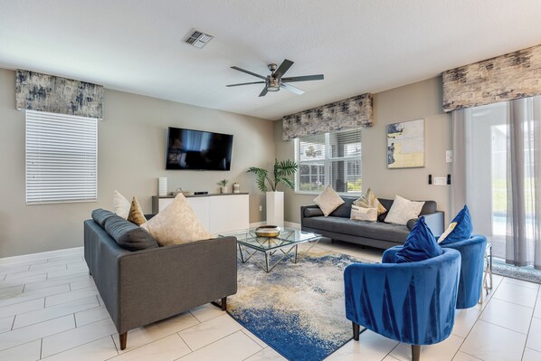 A modern living room with a grey sofa, blue armchairs, a glass coffee table, and a ceiling fan.
