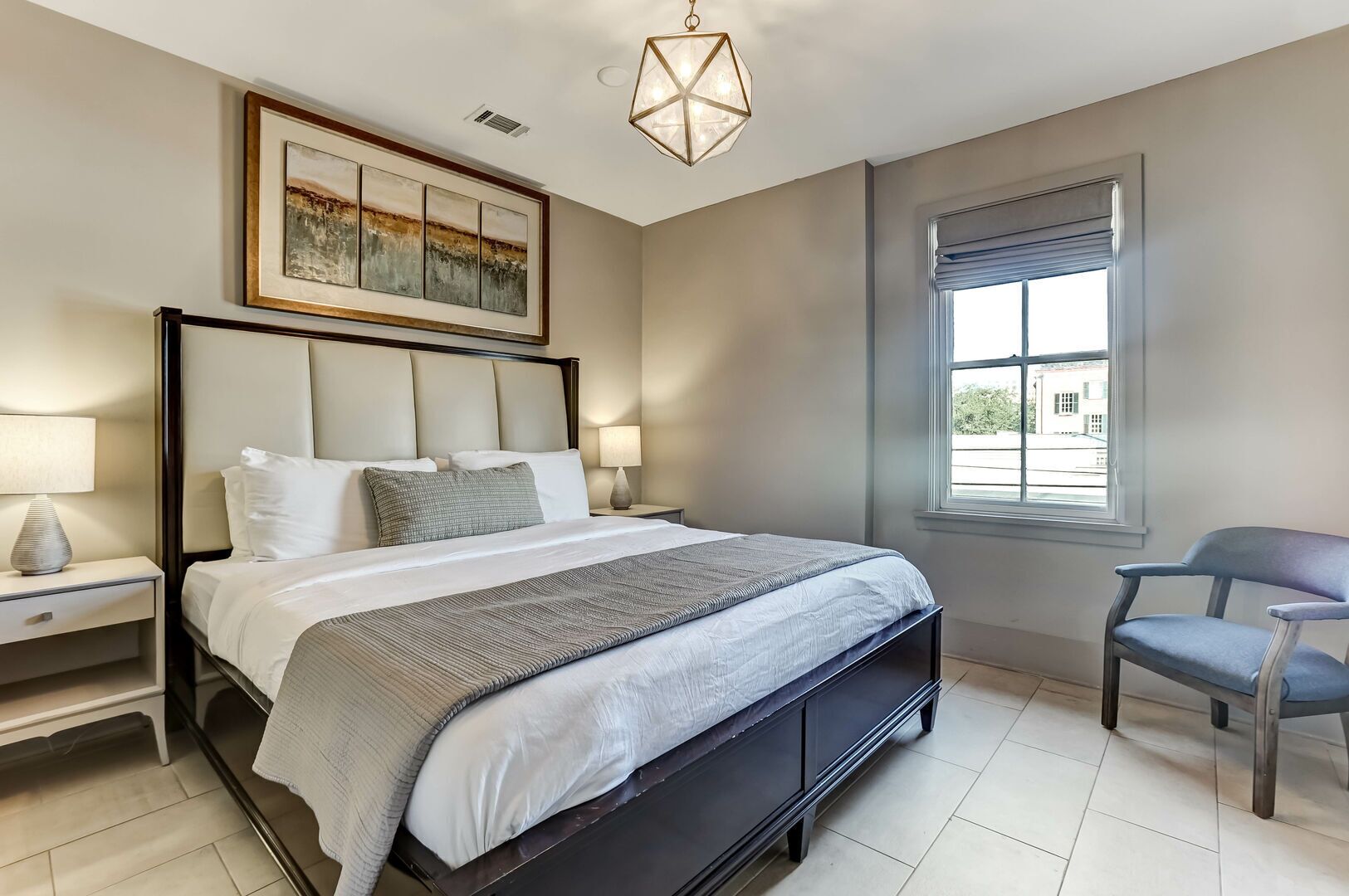 A bedroom with a large bed, bedside lamps, a chair, a window, and artwork on the wall.