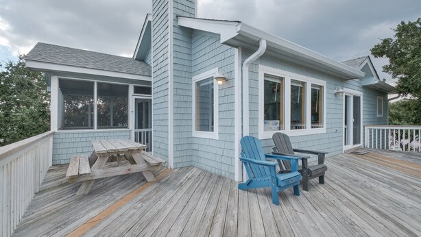A covered deck with a picnic table and Adirondack chairs, a house with light blue siding and white trim, and a cloudy sky. - 1747 - Resting Beach Place, Pool, Hot Tub & Game Room | Sleeps 15 in Corolla (Corolla)