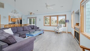 A spacious living room with a large sectional sofa, a glass coffee table, and a fireplace. There are multiple windows providing natural light and a view of the outdoors. - 1747 - Resting Beach Place, Pool, Hot Tub & Game Room | Sleeps 15 in Corolla (Corolla)
