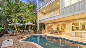 Un espace piscine avec des chaises longues et des parasols, une terrasse en bois et un bâtiment à deux étages doté d’un balcon.