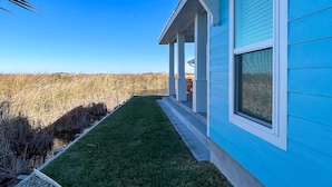 A house with a blue exterior, a well-maintained lawn, and a view of a natural landscape. - Golf Cart Included, Shared Pool, Hot Tub, Boardwalk to the Beach (Port Aransas)