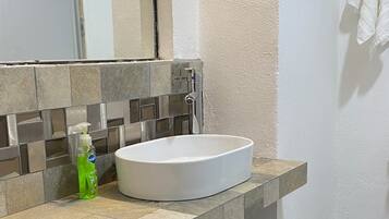 Un baño con lavamanos blanco, espejo, dispensador de jabón y cubo de basura.