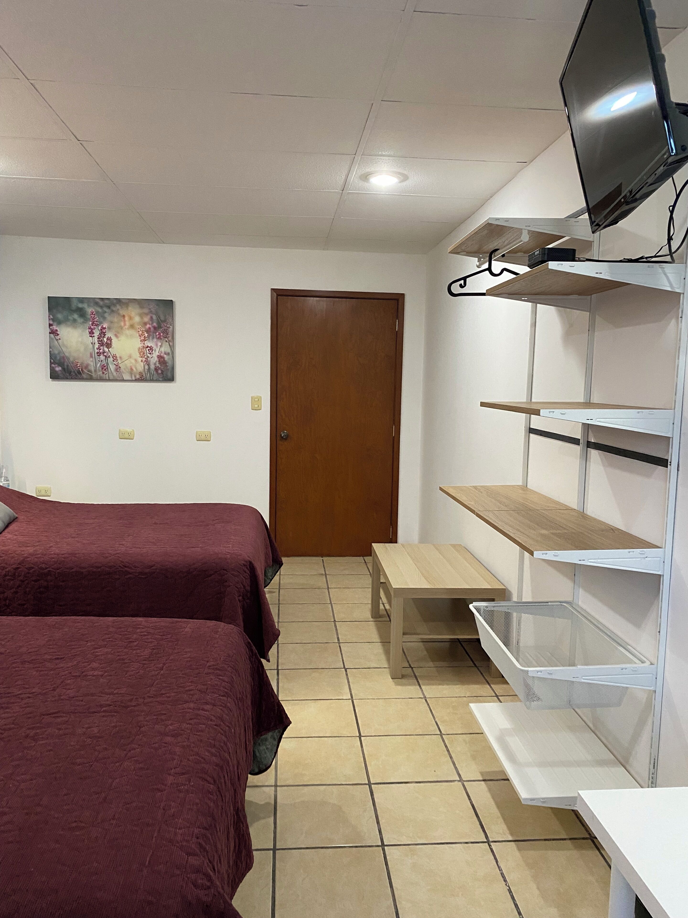 A room with two beds, a wooden door, a small table, and a TV mounted on the wall.