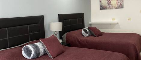A hotel room with two beds, each with a headboard and a pillow. There is a nightstand with a lamp, a wall-mounted shelf, and a painting on the wall.
