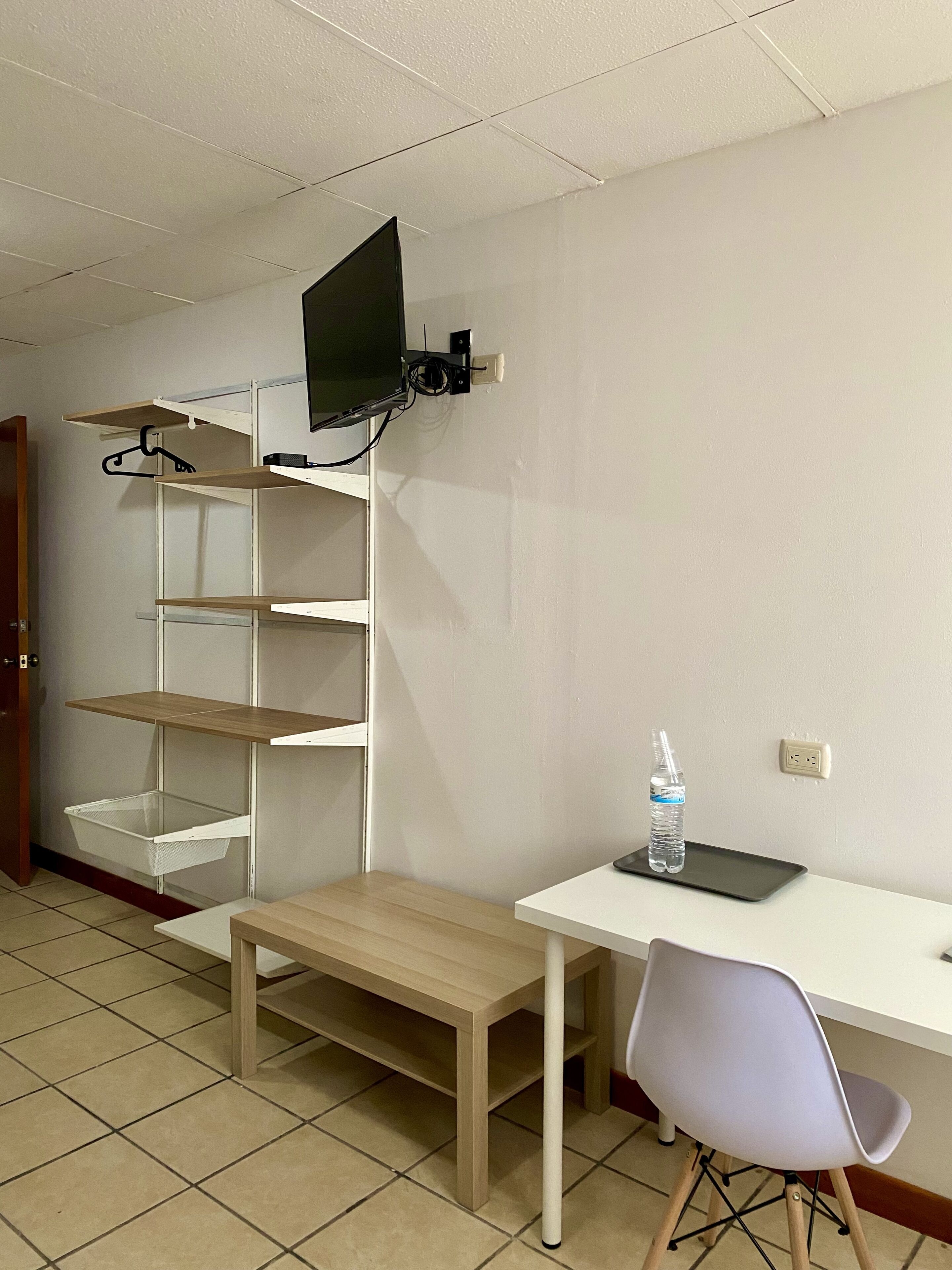 A room with a white desk, a chair, a TV mounted on a shelf, and a bottle on the desk.