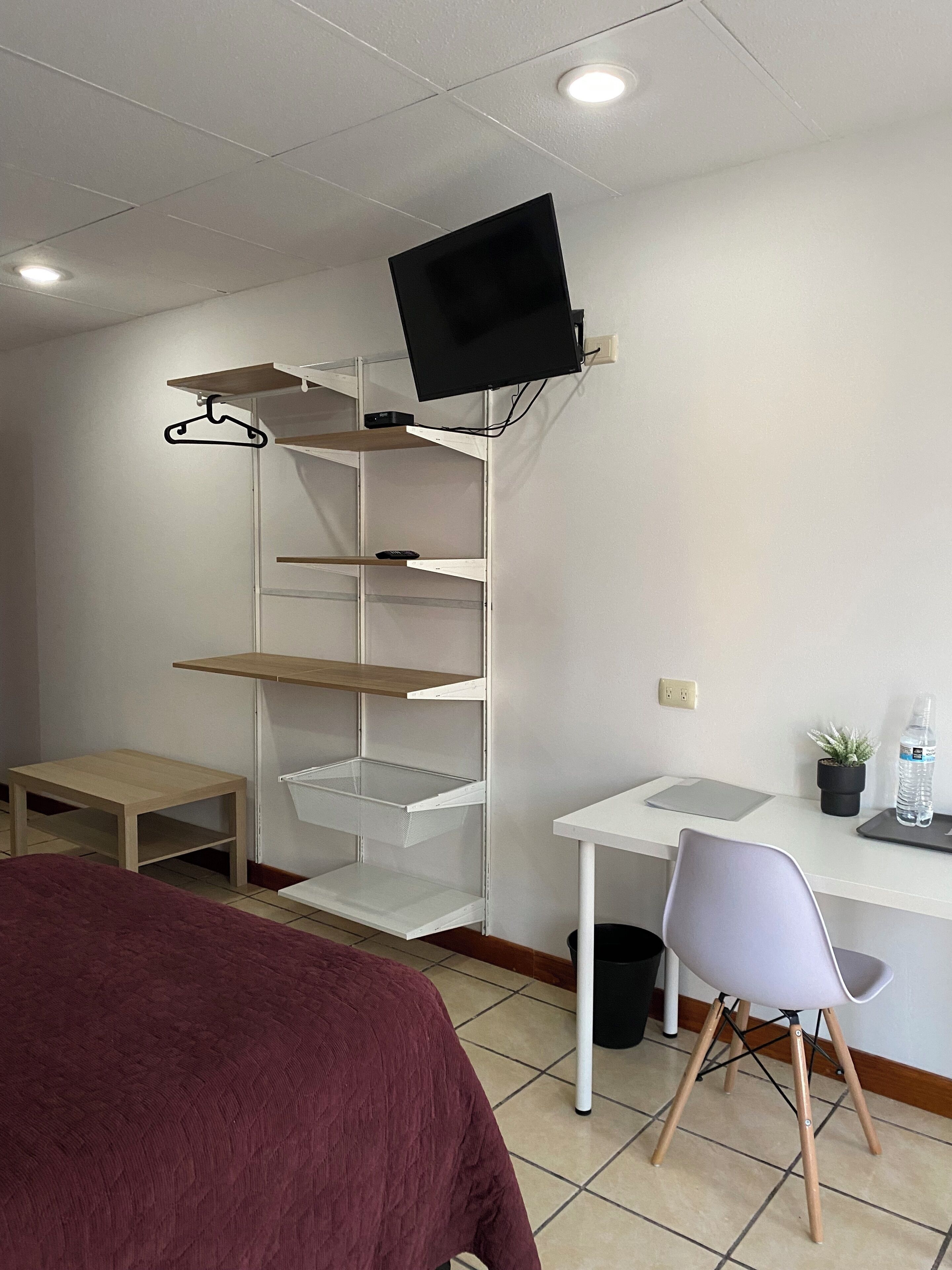 Une pièce comprenant un lit, un bureau, une chaise, un téléviseur fixé à un mur blanc et une étagère contenant divers objets.