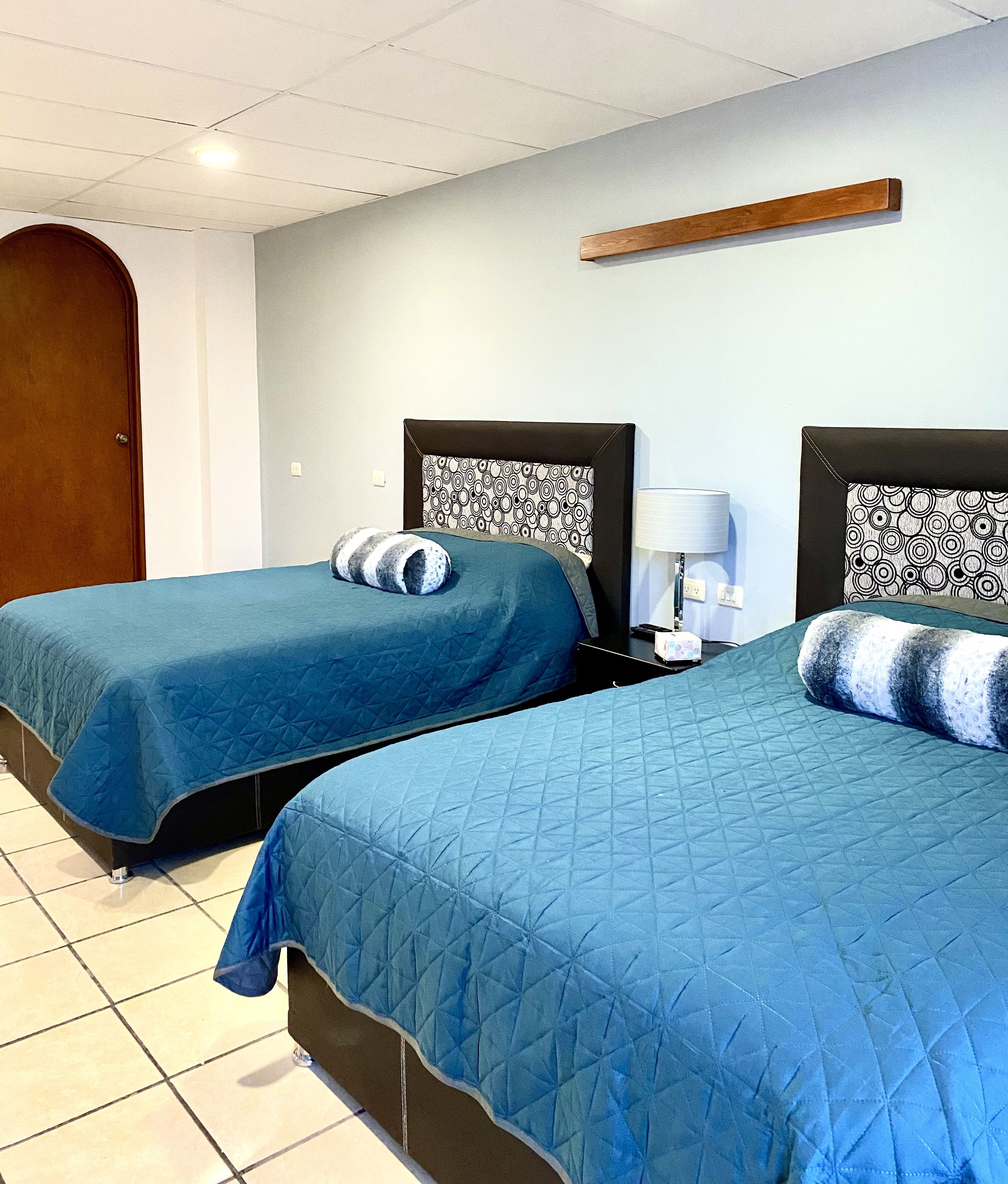 A hotel room with two beds, each with a blue quilted bedspread and matching pillows. There is a wooden headboard on each bed, and a small table with a lamp between them.