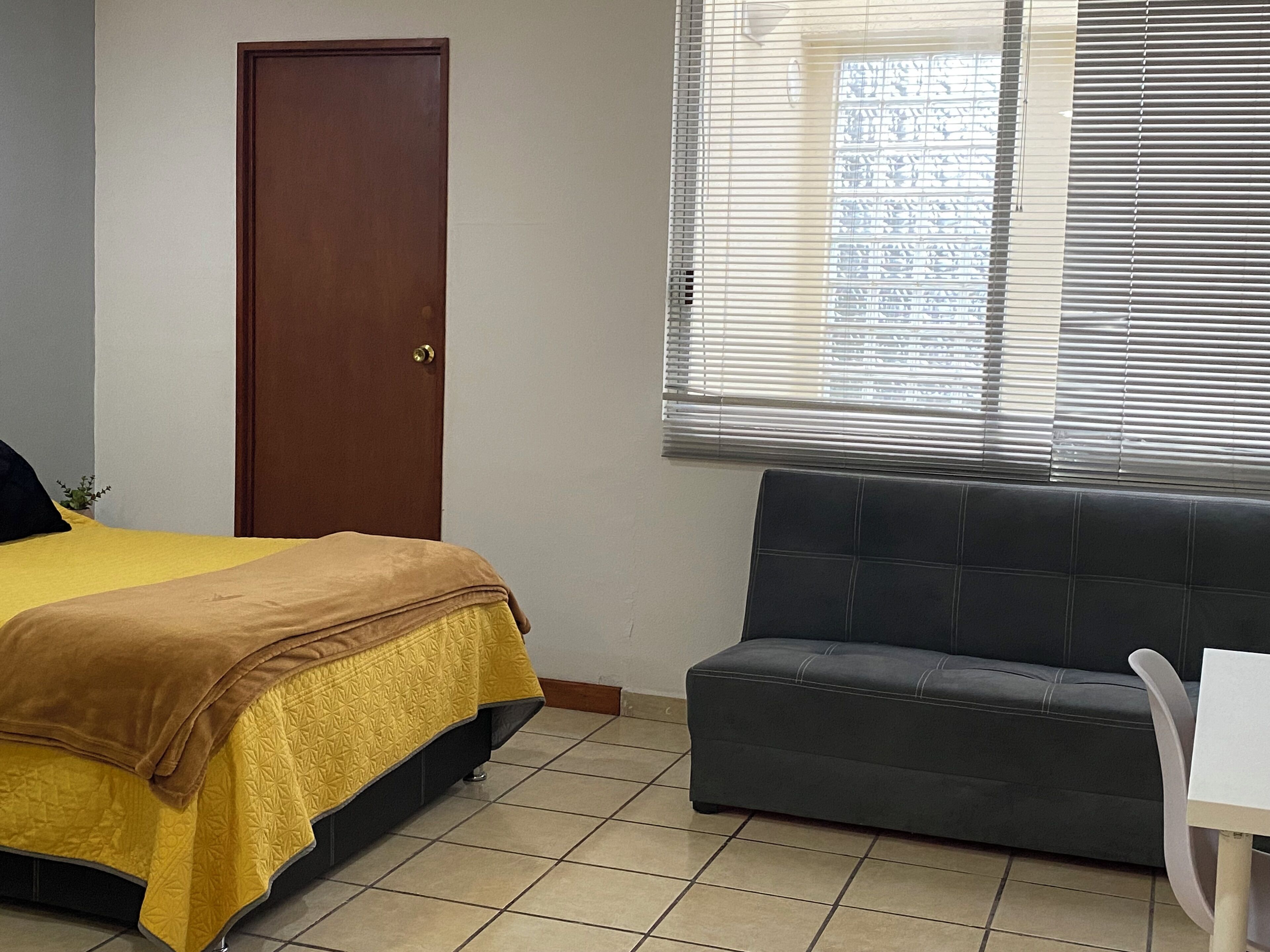 A room with a bed, a sofa, a door, and a window with blinds.