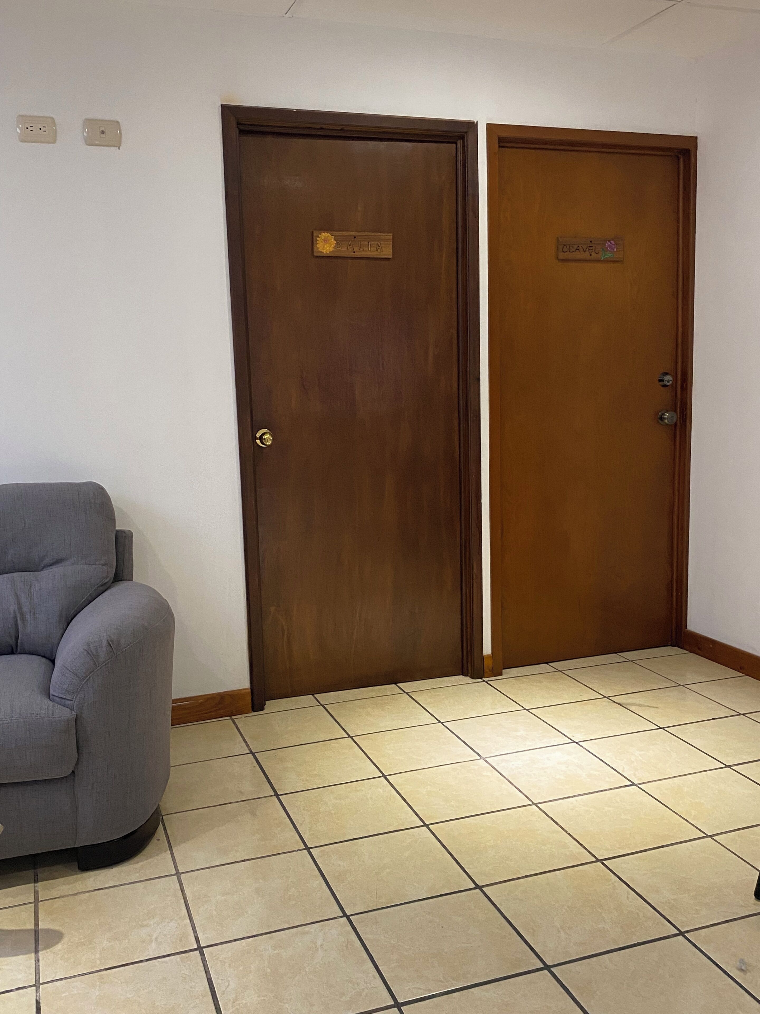 A room with two wooden doors, one labeled "DALLA" and the other "CEAVEL," a grey upholstered armchair, and a tiled floor.