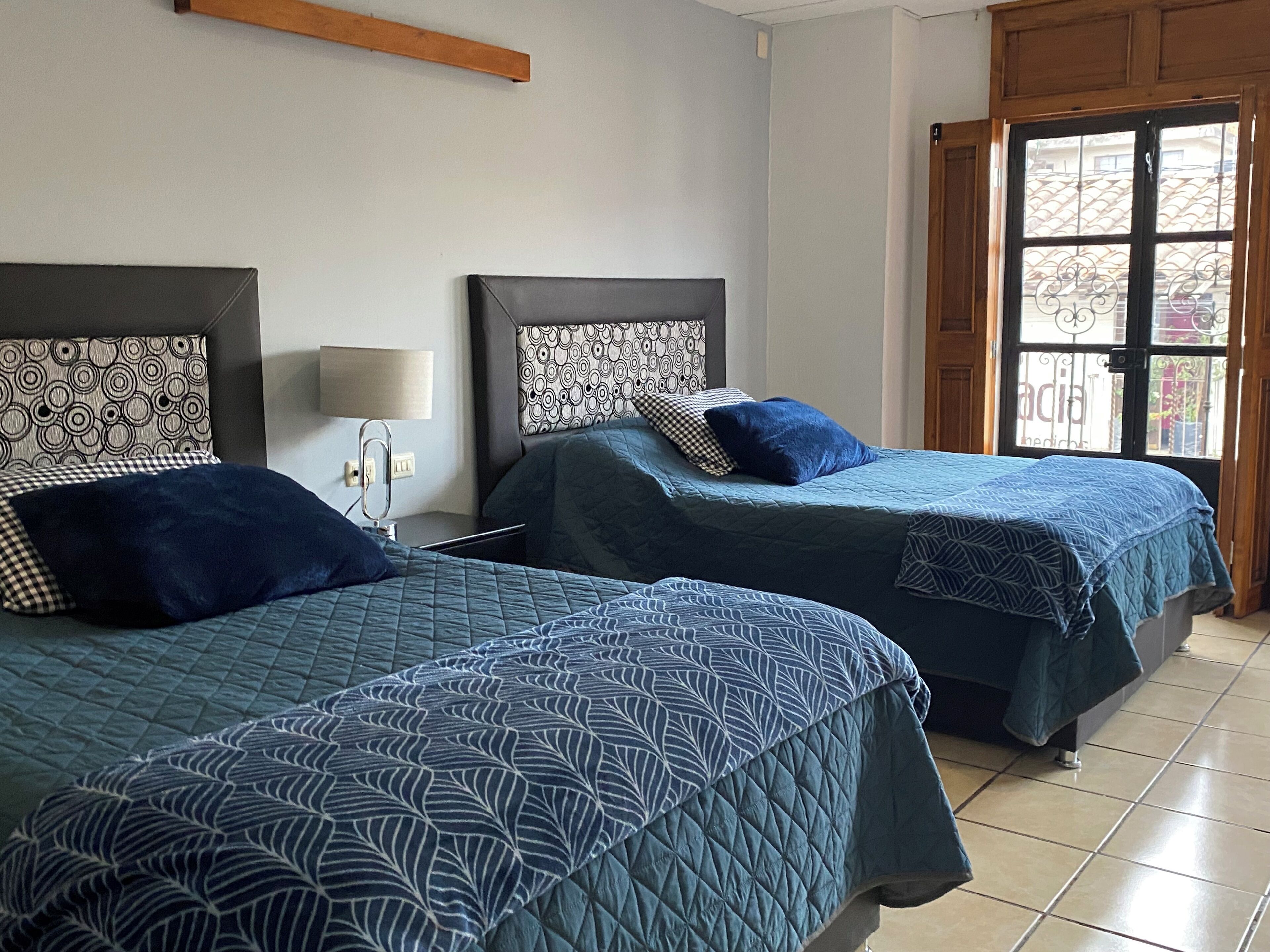 Two single beds with patterned bedding, a bedside table with a lamp, and a window with a view of buildings.