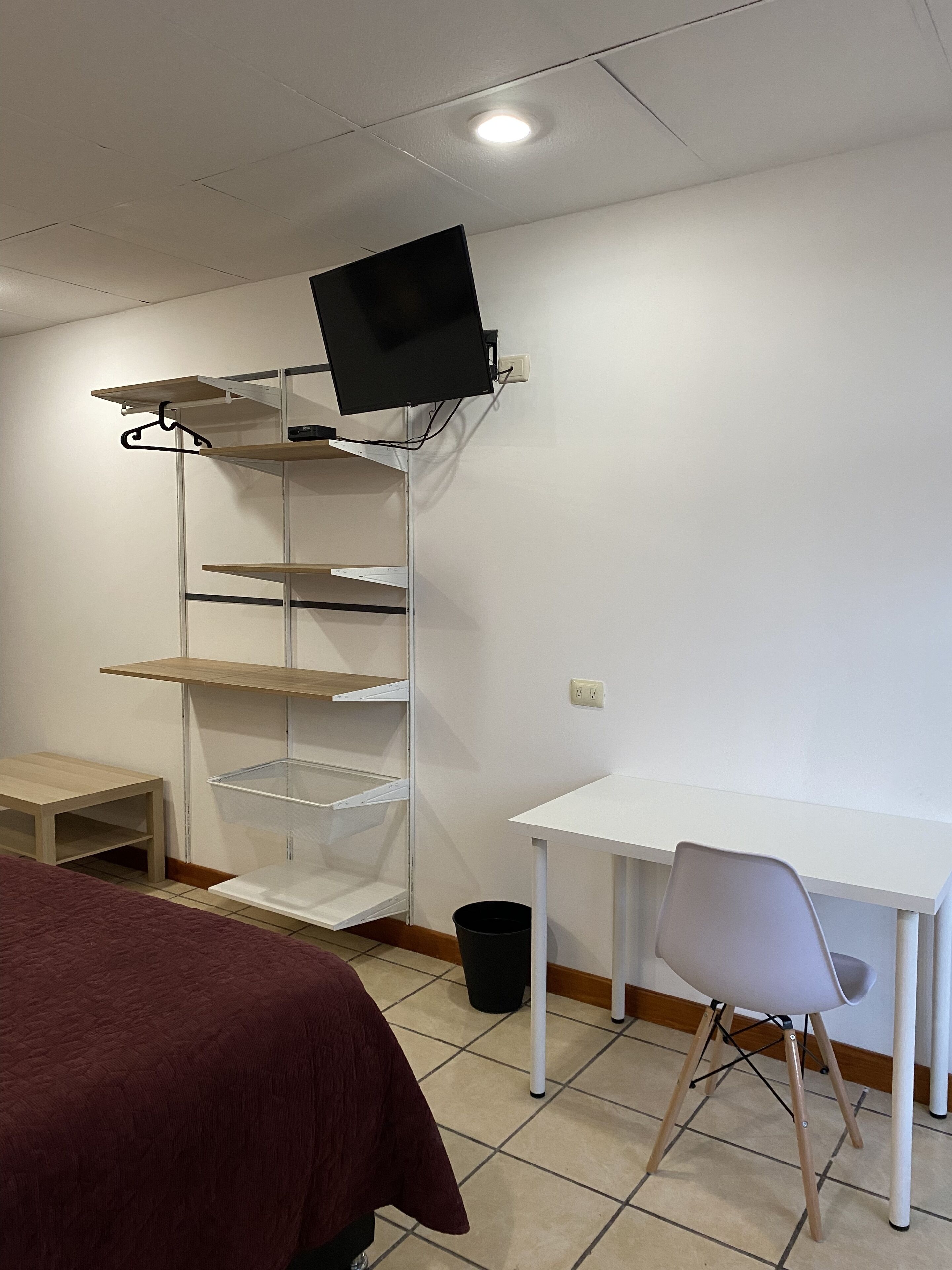 Ein Zimmer mit weißem Schreibtisch, einem Stuhl, einem Bett mit roter Decke und einem Schrank mit Fernseher.