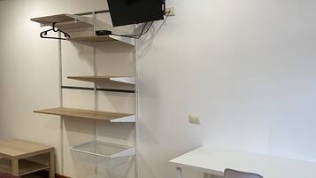 A room with a white desk, a chair, a bed with a red blanket, and a shelving unit with a TV.