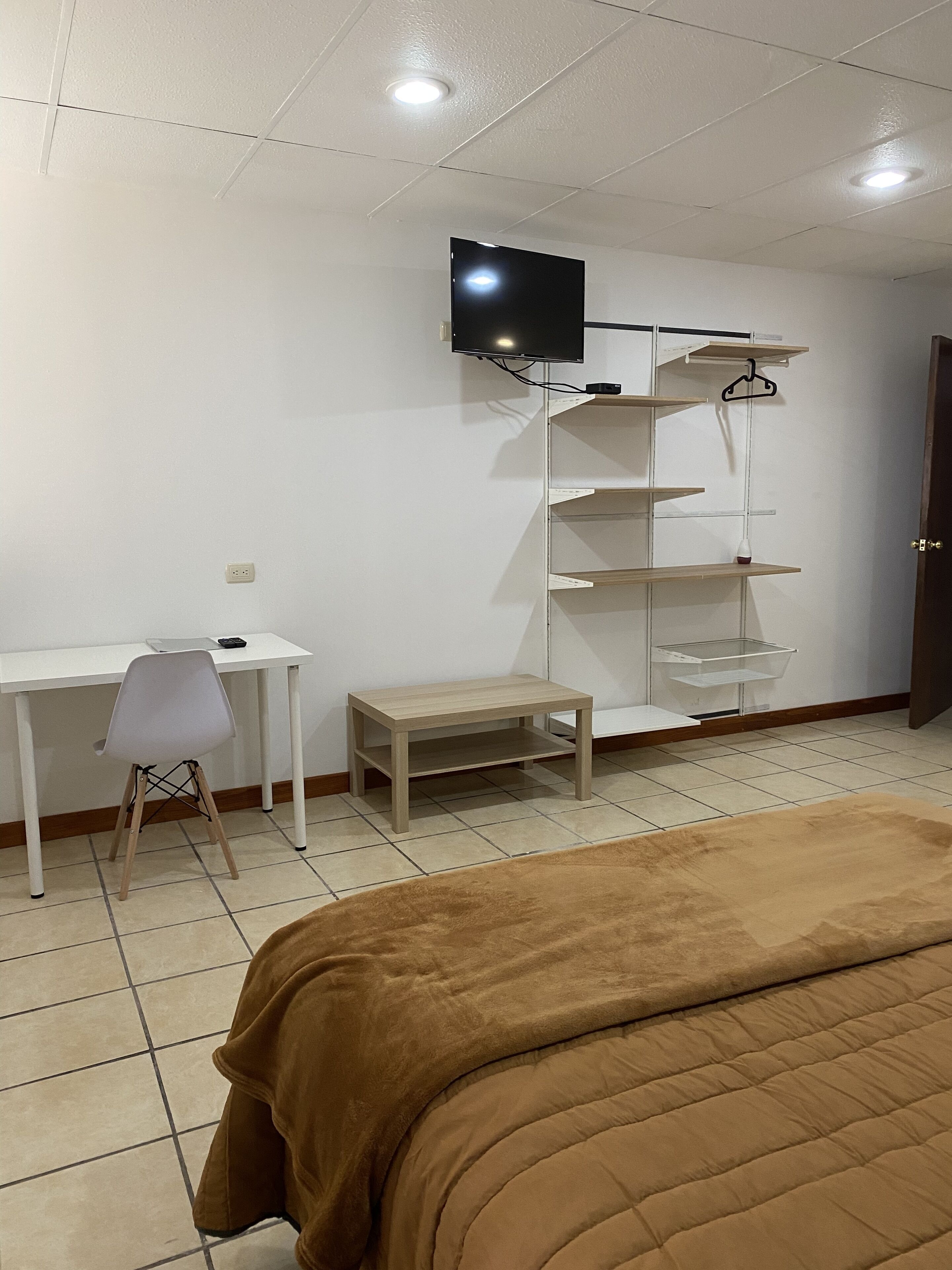 A bedroom with a bed, a desk, a chair, a TV, and shelves.