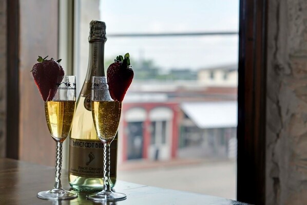 Dos copas de champán con fresas y una botella de Barefoot Bud Champagne sobre una mesa con vistas a un puente y edificios.