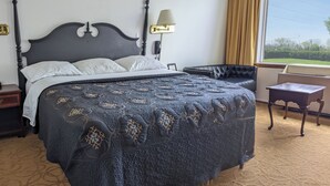 A neatly made bed with a dark quilt, a bedside table, a lamp, and a view of the outdoors through a window. - South Side Inn - Burlington (South Burlington)