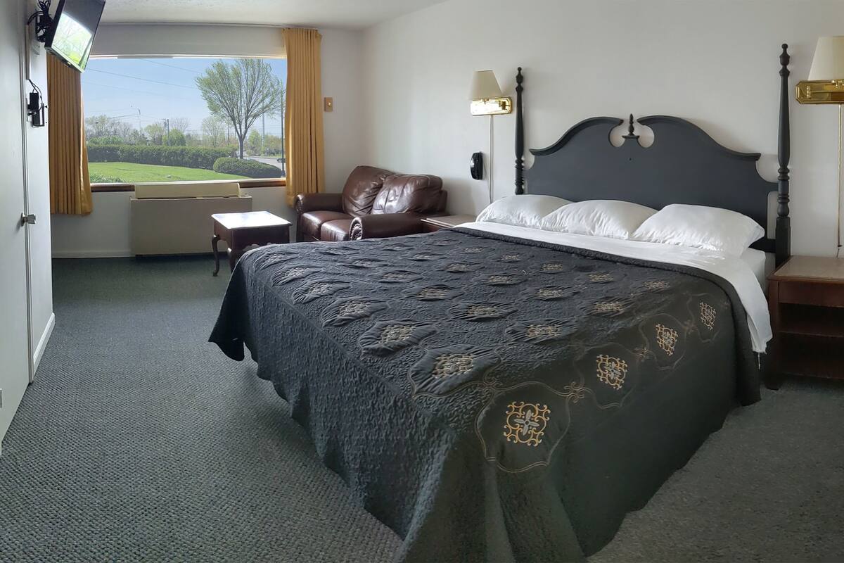 Un cuarto de hotel con una cama grande, un sillón de cuero, un escritorio y vistas a un paisaje verde a través del ventanal.