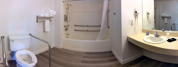 A bathroom with a toilet, a shower with grab bars, a sink, and a hairdryer. - South Side Inn - Burlington (South Burlington)