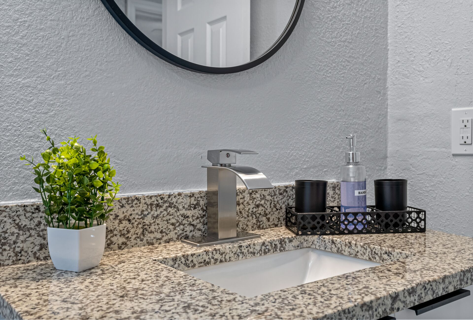 Casa de banho com bancada de granito, lavatório, espelho, planta num vaso e dispensador de álcool em gel.
