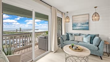 A balcony with a view of the ocean and a pier, a grey sofa with cushions, a round coffee table with decorative items, and two pendant lights.