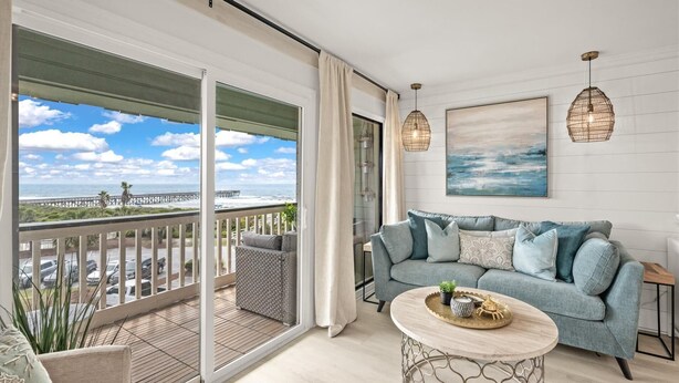 A balcony with a view of the ocean and a pier, a grey sofa with cushions, a round coffee table with decorative items, and two pendant lights.