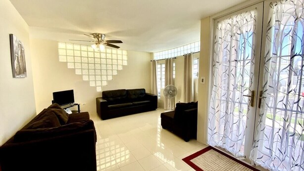 A living room with a sofa, a television, a ceiling fan, and a sliding glass door with decorative curtains. - Dorado Vacation Home-Wi-Fi & AC (Dorado)